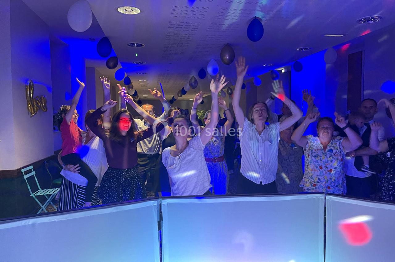Des personnes dansent et lèvent les bras dans une salle éclairée par des lumières colorées, décorée de ballons.