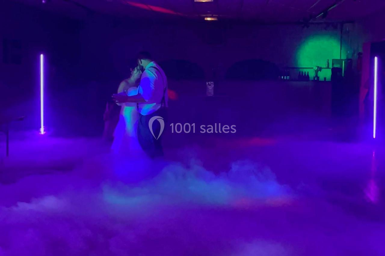 Un couple danse dans une salle sombre éclairée par des lumières violettes et vertes, entouré d'une brume au sol.