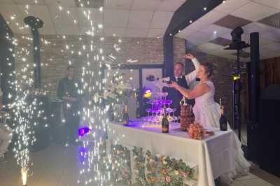 Un couple pose près d'une table décorée de fleurs et d'une pyramide de coupes de champagne dans une salle éclairée.