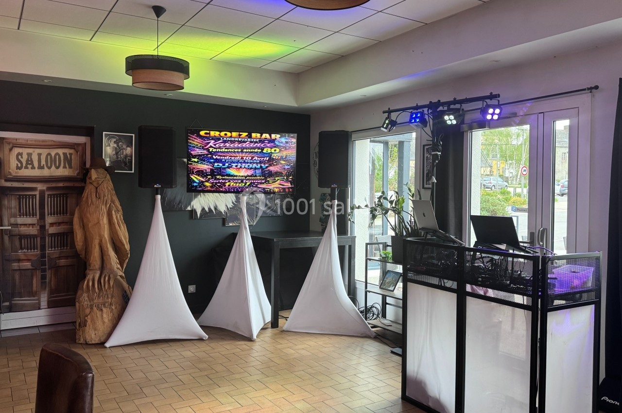 Salle équipée pour un événement avec éclairage coloré, écran affichant une affiche, et espace DJ aménagé.