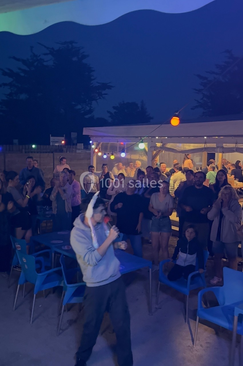 Un homme chante au micro devant un public assis et debout, éclairé par des guirlandes lumineuses en soirée.
