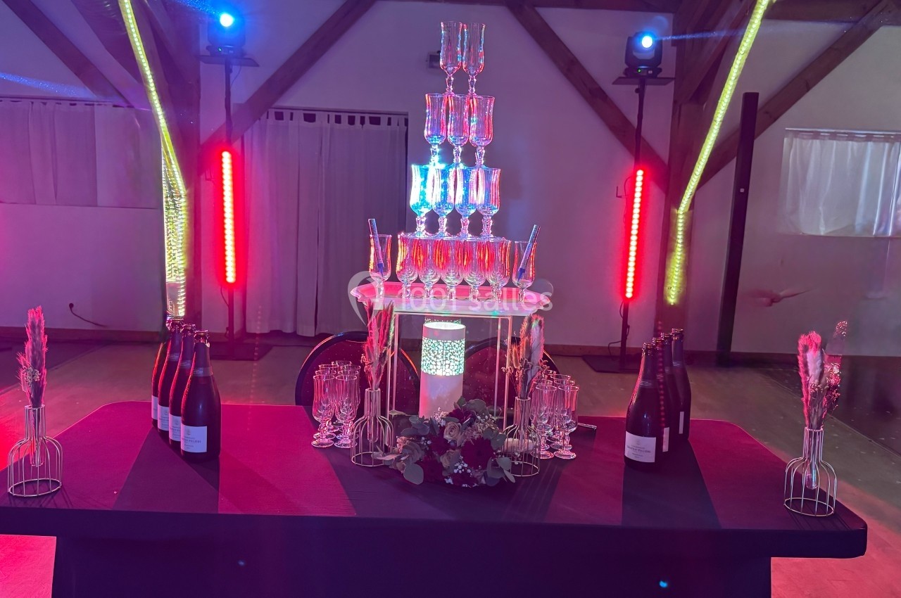 Tour de coupes en verre empilées sur une table décorée, entourée de bouteilles et éclairée par des lumières colorées.