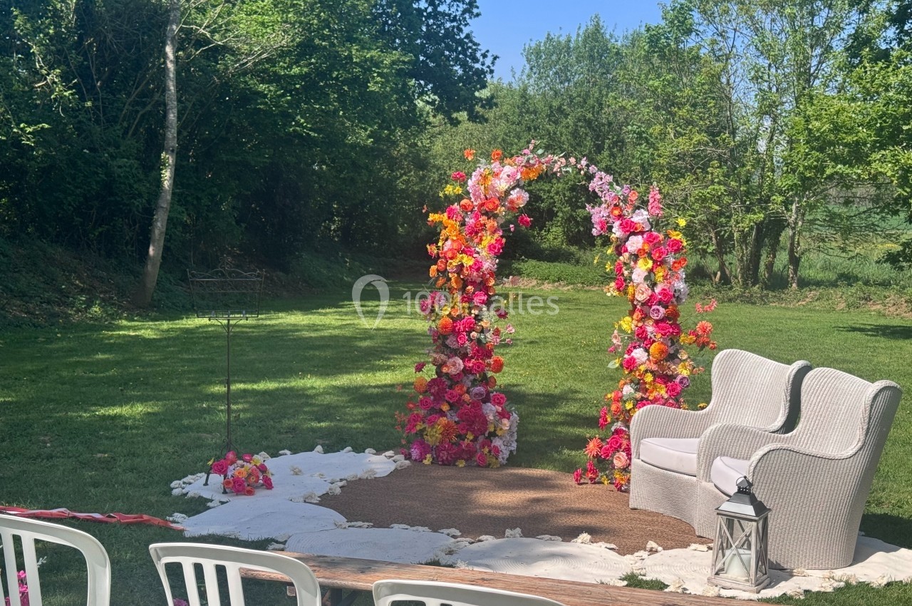 Arche florale colorée avec fauteuils en osier, installée en extérieur sur une pelouse entourée d'arbres.