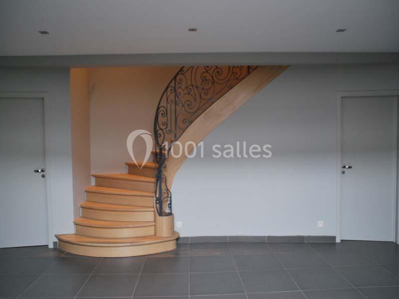 Location salle Ciney (Province de Namur) - Le Domaine d'Achêne #9 Escalier en bois avec rampe en fer forgé dans une pièce aux murs clairs et sol en carrelage gris.