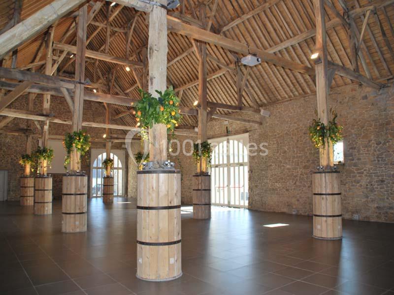 Location salle Ciney (Province de Namur) - Le Domaine d'Achêne #12 Salle spacieuse avec charpente en bois apparente, piliers décorés de plantes et murs en pierre.