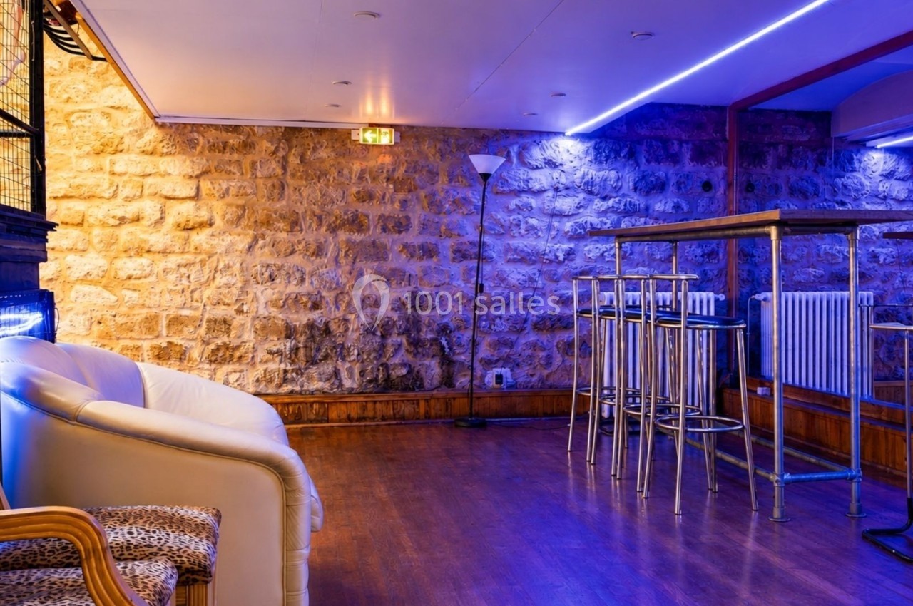 Salle avec murs en pierre, éclairage bleu, tables hautes en métal et fauteuils, sol en parquet.