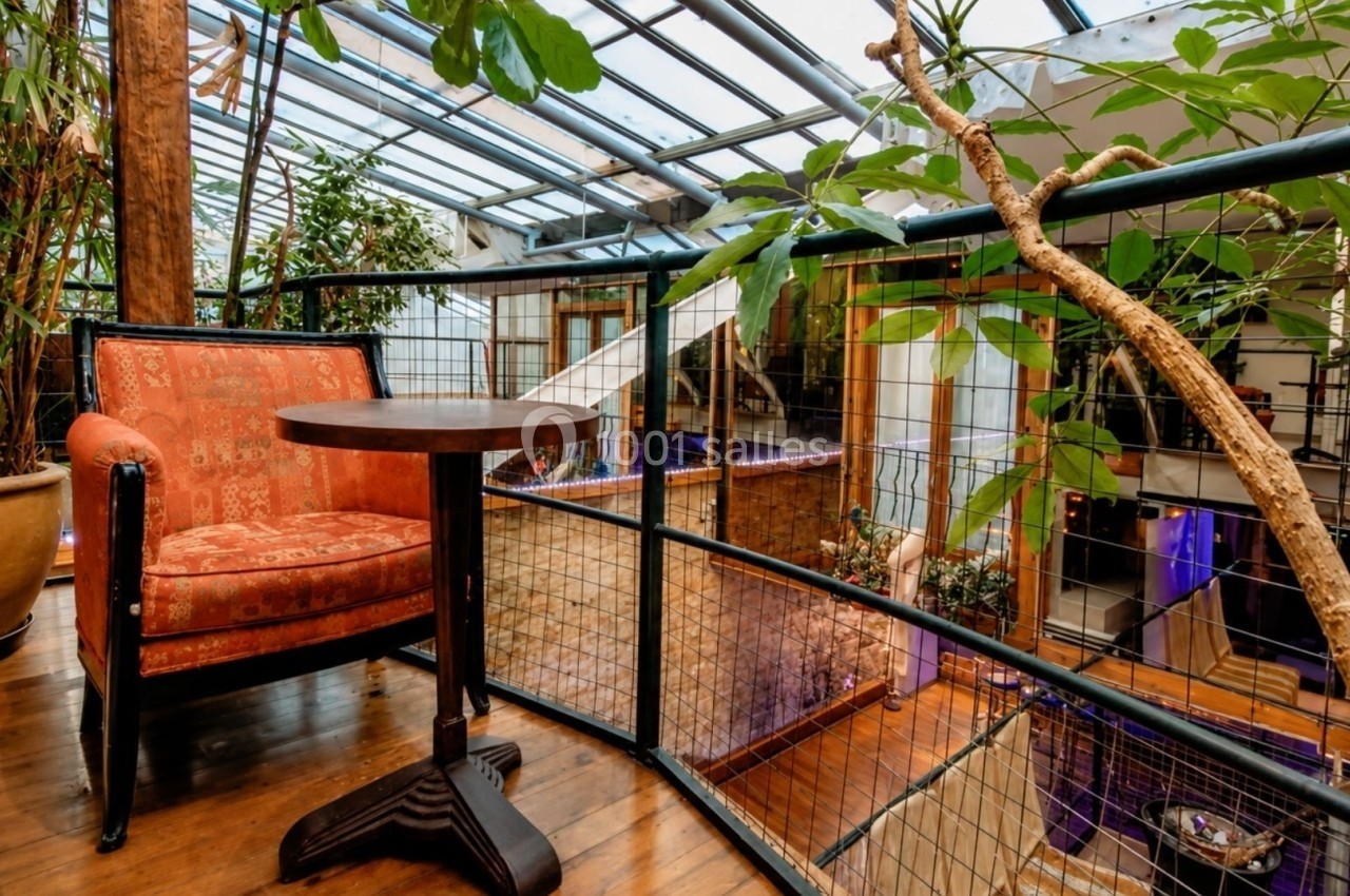 Fauteuil orange et table ronde en bois dans un espace lumineux avec verrière et plantes vertes.