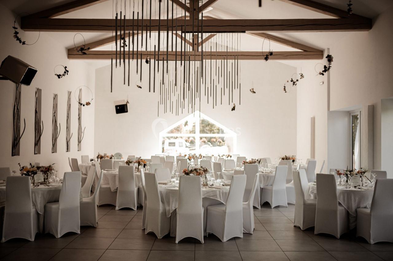 Salle de réception lumineuse avec tables rondes dressées, chaises blanches et décorations suspendues au plafond.