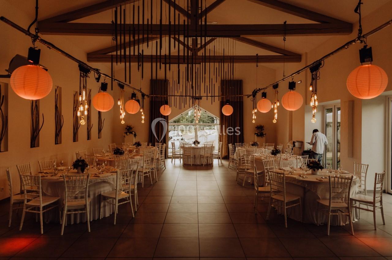 Salle de réception décorée avec des lanternes suspendues, des tables rondes dressées et des chaises blanches.