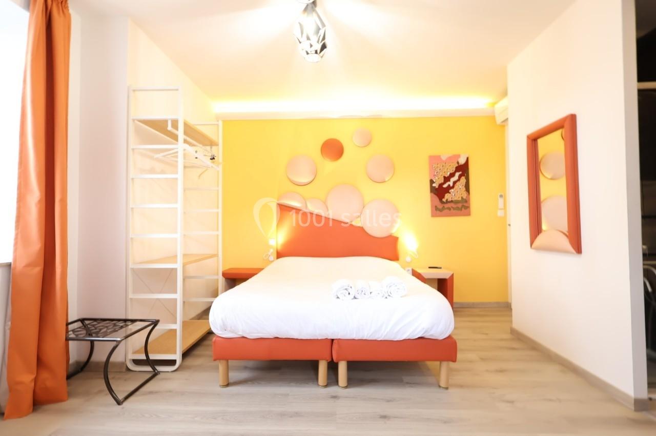 Chambre lumineuse avec lit double, mur jaune décoré, étagère ouverte, rideaux orange et sol en bois clair.