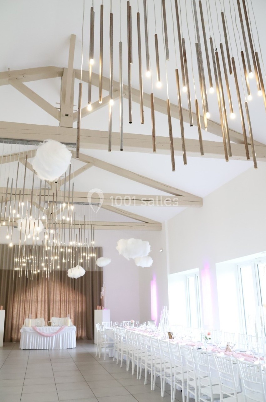 Salle de réception lumineuse avec des tables décorées, des chaises blanches et des suspensions modernes en bois.