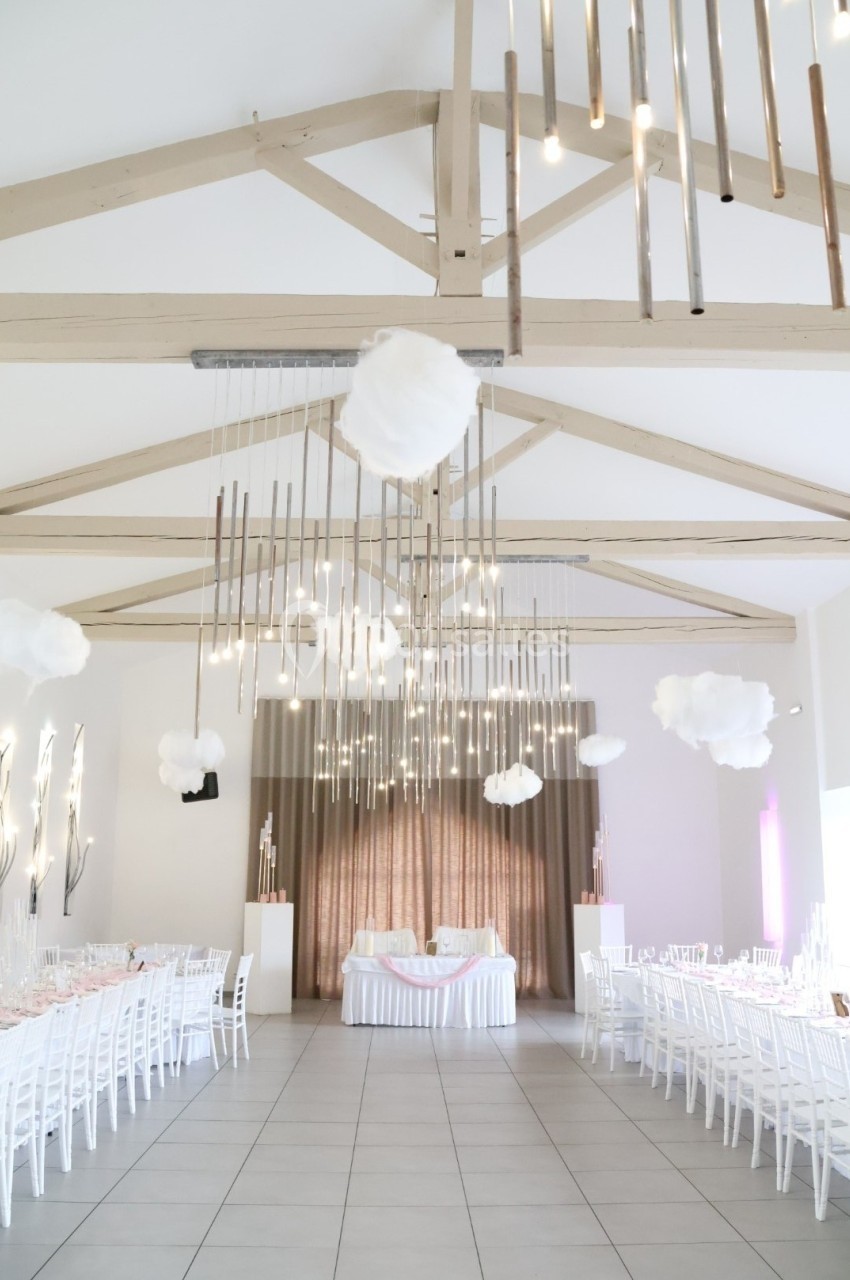Salle de réception lumineuse avec tables blanches alignées, décorations suspendues et poutres apparentes en bois clair.