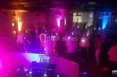 Salle de réception décorée avec des tables rondes, nappes blanches, chaises ornées de nœuds beige et éclairage vert.