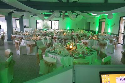 Salle de réception décorée avec des tables rondes, nappes blanches, chaises ornées de nœuds beige et éclairage vert.
