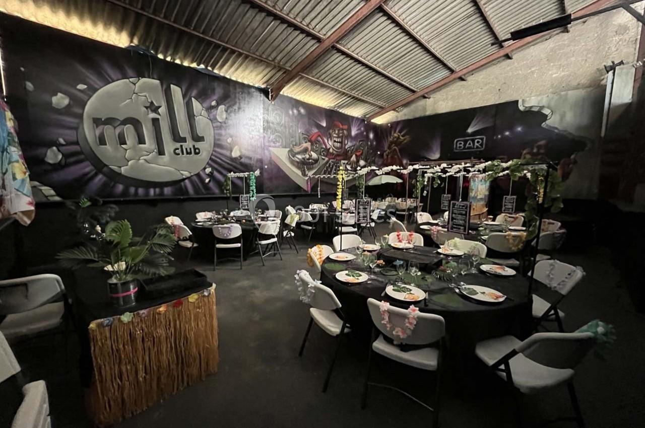 Salle décorée pour un événement avec des tables rondes dressées, des éléments tropicaux et un mur avec le logo ’Milli Club’.
