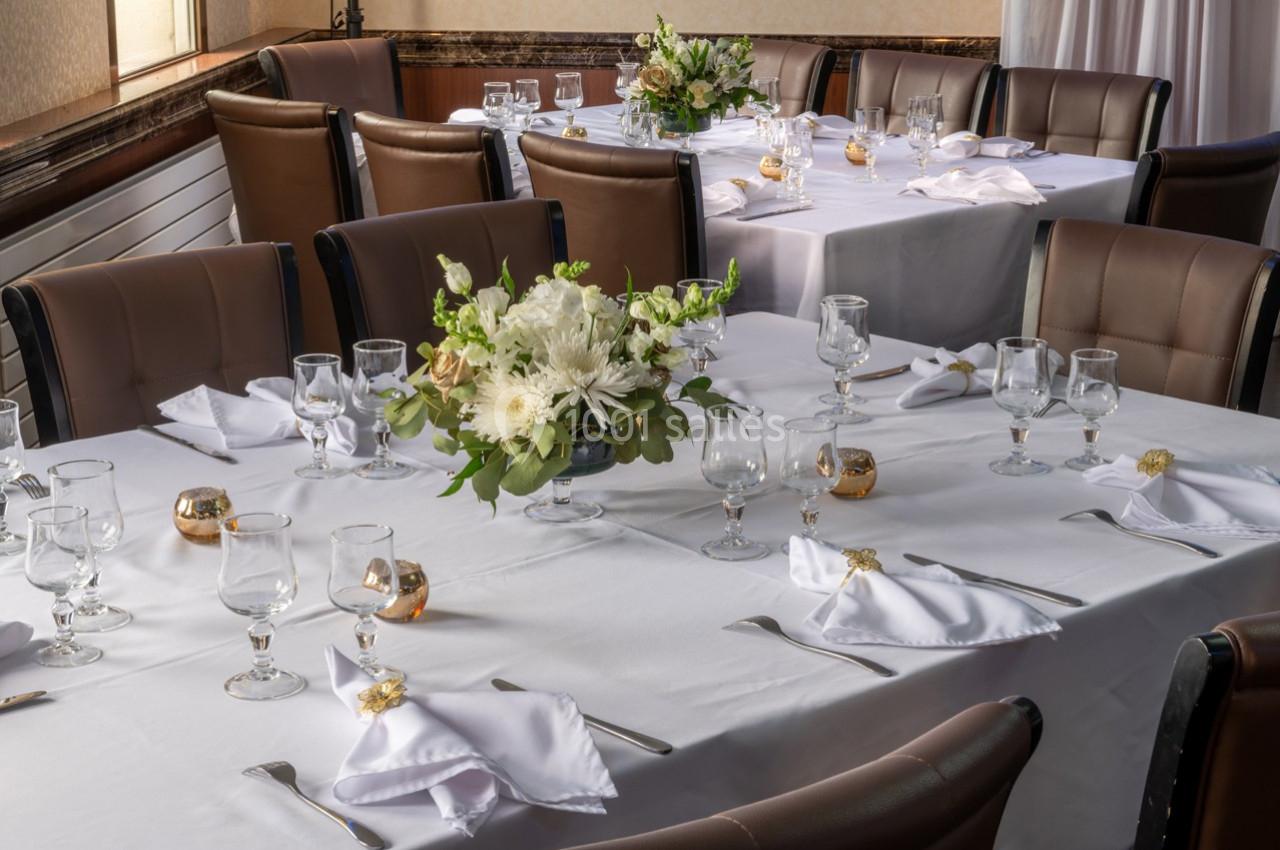 Salle de réception avec tables dressées, nappes blanches, couverts, verres et centre de table floral.
