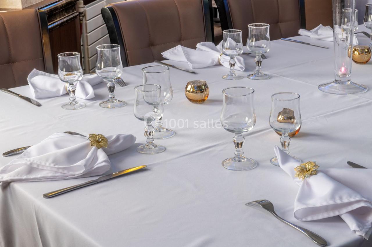 Table dressée avec nappes blanches, serviettes pliées, verres, couverts et petites décorations dorées.