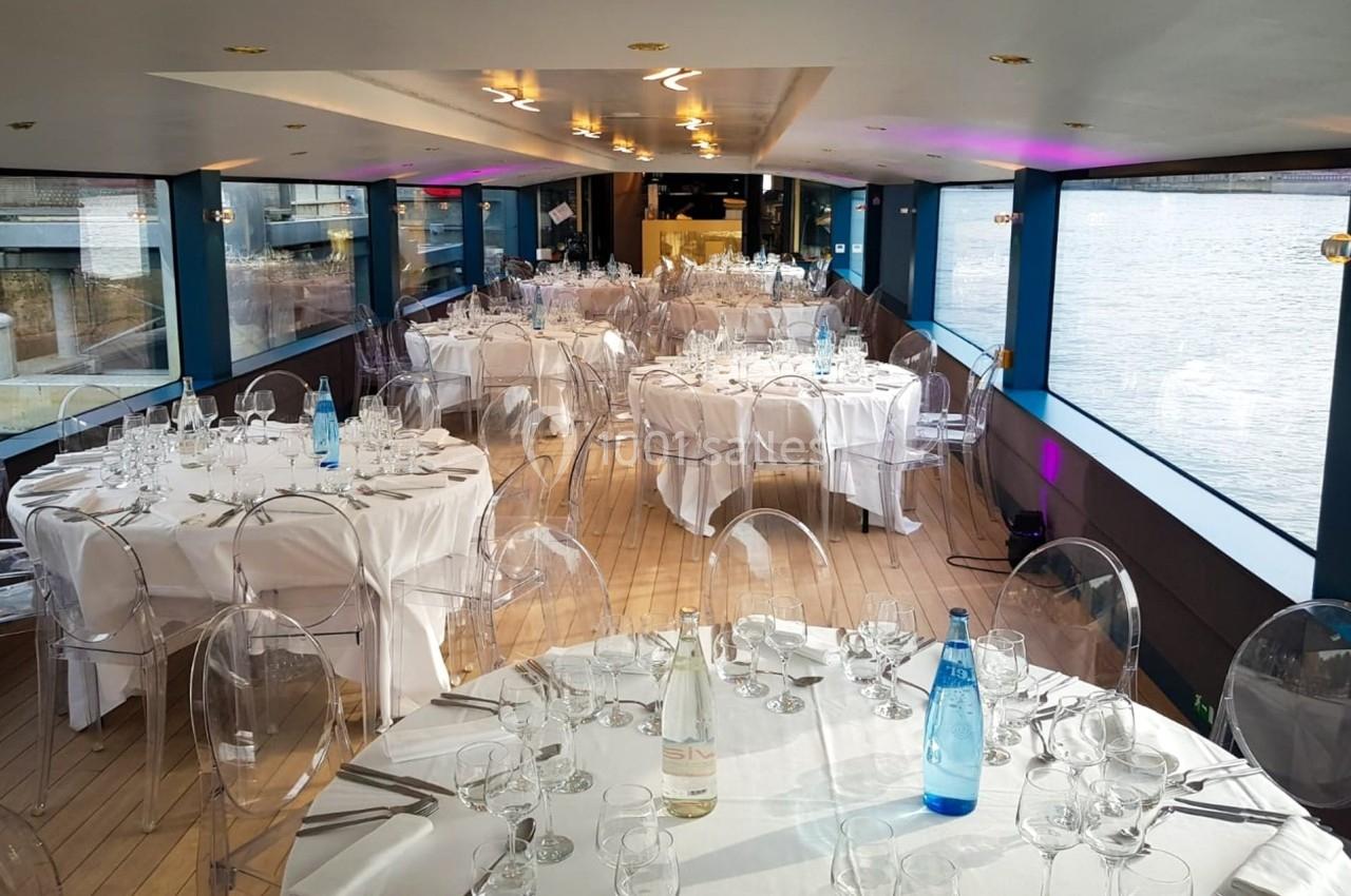 Salle de réception sur un bateau avec tables rondes dressées, nappes blanches et chaises transparentes, vue sur l'eau.