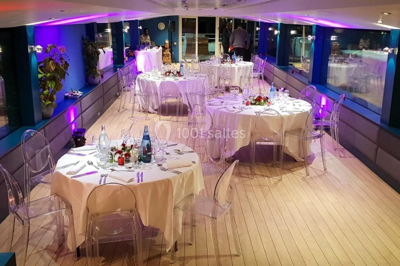 Salle de réception élégante sur un bateau, avec tables rondes dressées, chaises transparentes et éclairage tamisé.