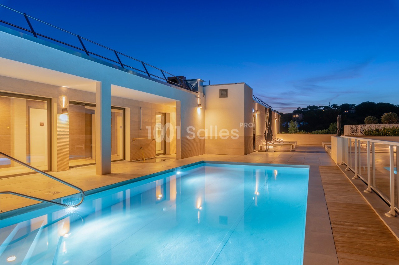 Piscine éclairée entourée d'une terrasse en bois et d'une maison moderne au crépuscule.