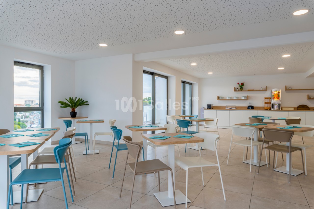 Salle lumineuse avec tables et chaises colorées, grandes fenêtres et coin cuisine en arrière-plan.