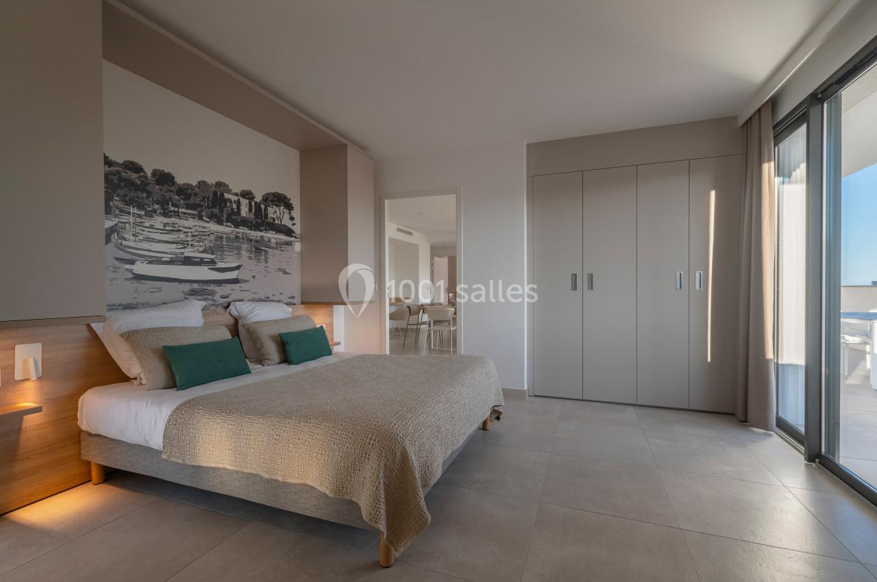 Chambre lumineuse avec lit double, décoration murale représentant un paysage, armoire intégrée et accès à un balcon.