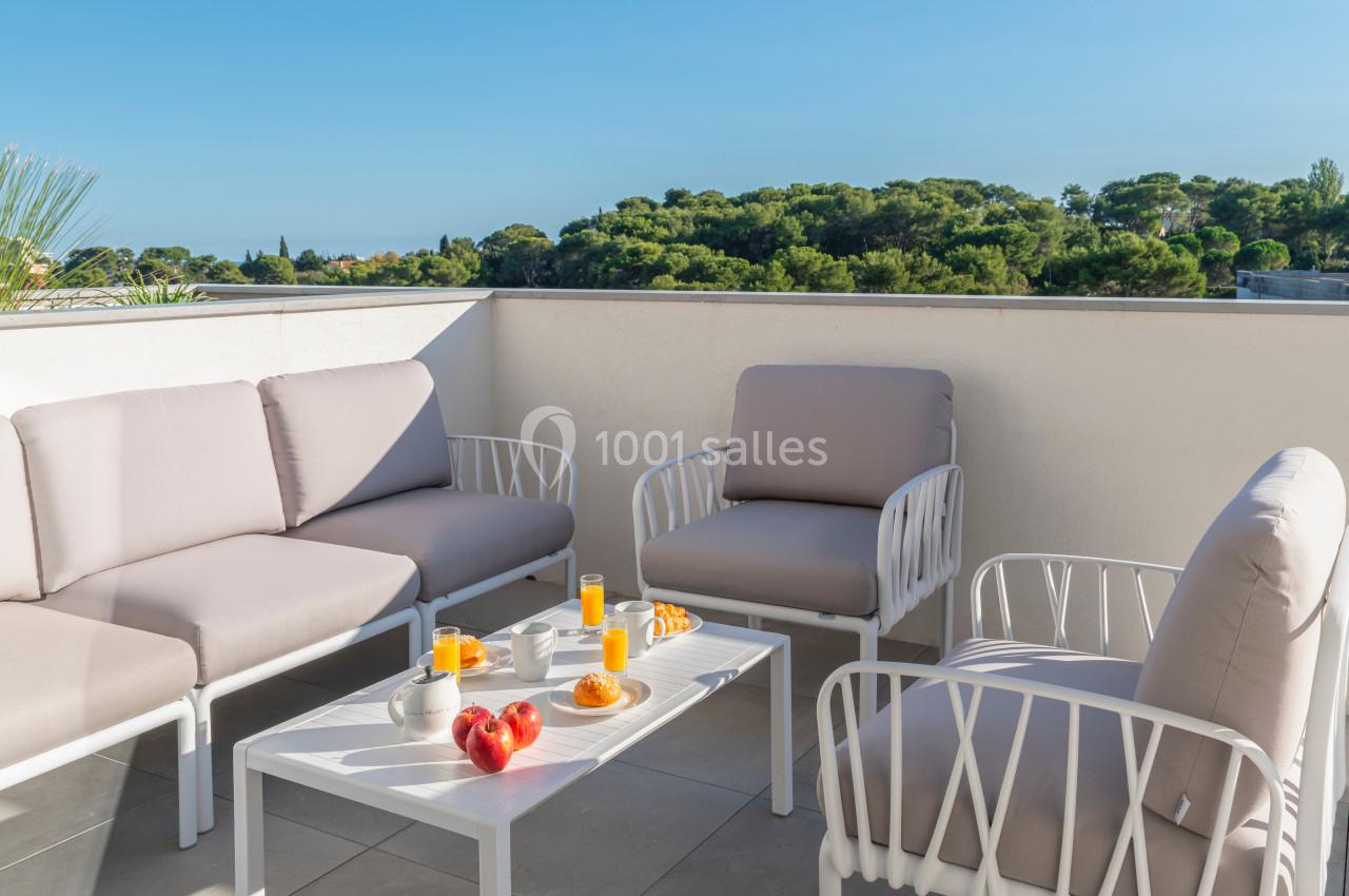 Salon de jardin avec canapés et table basse, petit-déjeuner servi, terrasse ensoleillée avec vue sur des arbres.