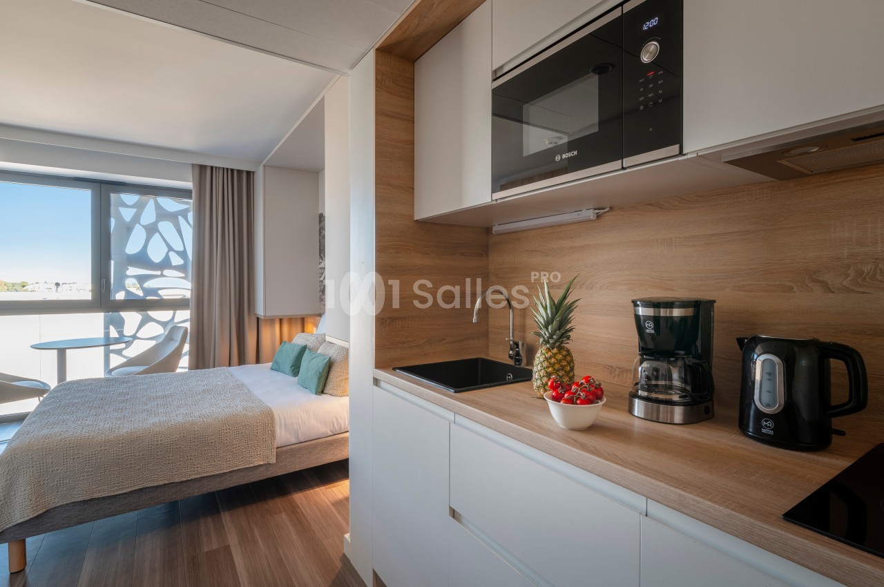 Cuisine équipée avec micro-ondes, cafetière, bouilloire et fruits frais, attenante à une chambre lumineuse avec lit double.