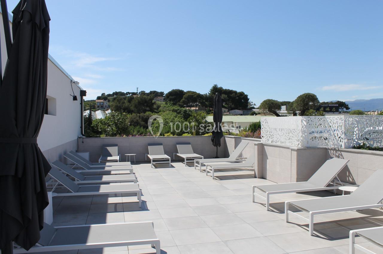 Terrasse ensoleillée avec transats gris, parasols noirs et vue sur un paysage verdoyant.