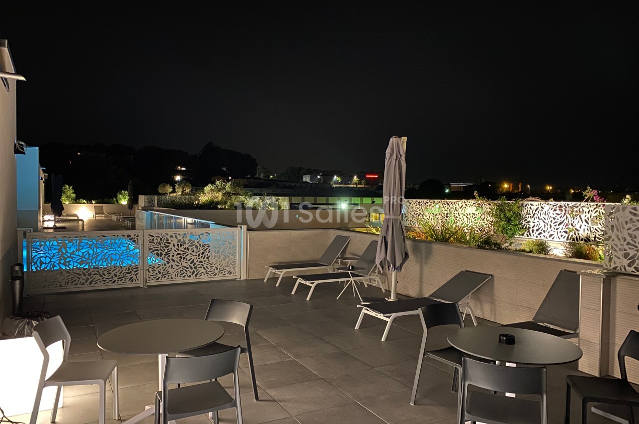 Terrasse extérieure éclairée la nuit avec des tables, des chaises longues et une vue sur une piscine illuminée.