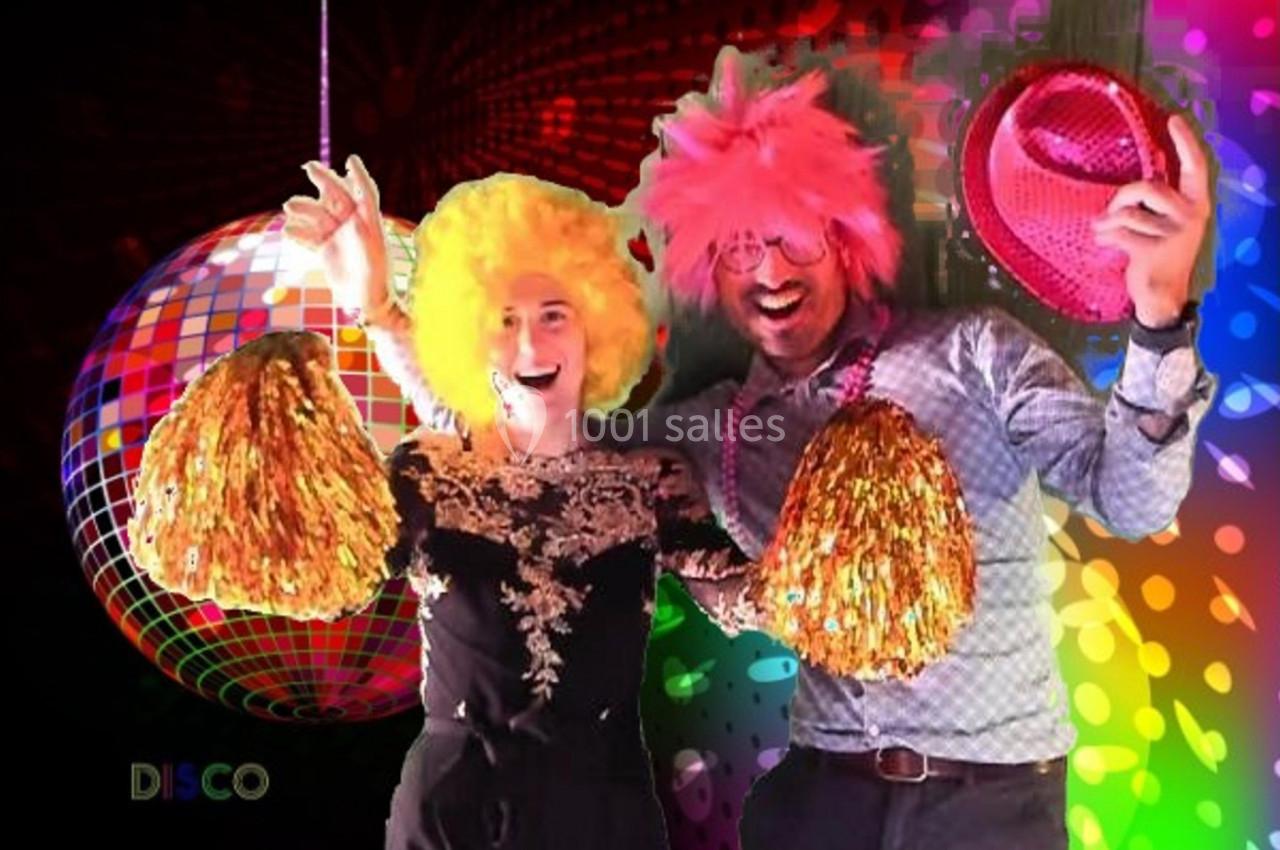 Deux personnes déguisées avec perruques colorées et accessoires festifs posent devant un décor disco lumineux.