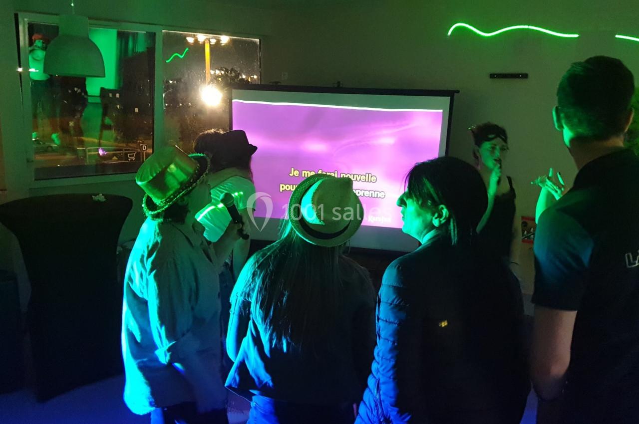 Des personnes chantent devant un écran de karaoké dans une pièce éclairée par des lumières colorées.