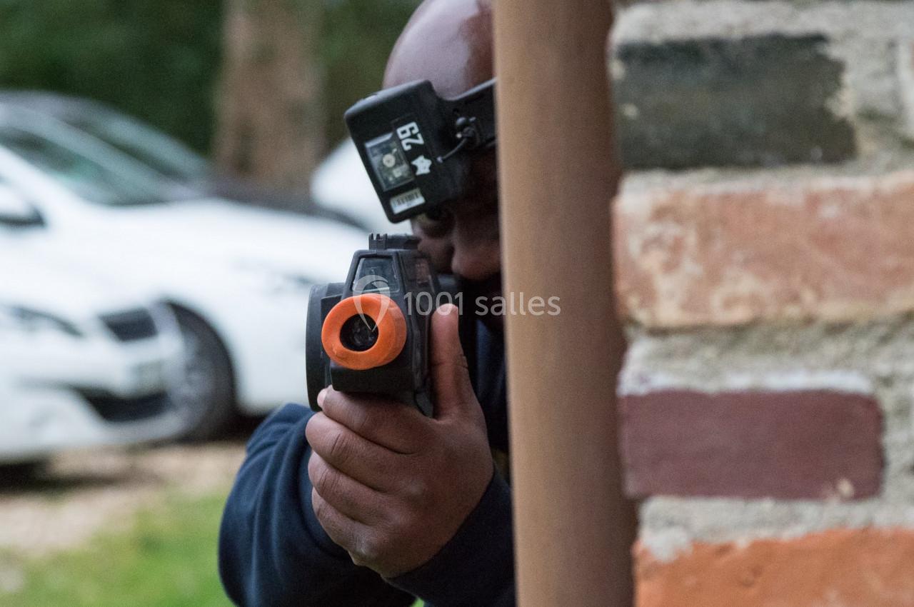 Homme tenant un pistolet laser, partiellement caché derrière un mur en briques, avec des voitures en arrière-plan.