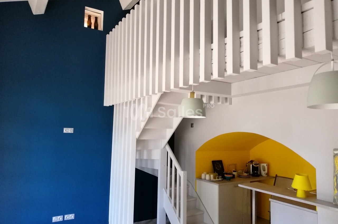 Escalier blanc moderne avec balustrade ajourée, mur bleu, coin cuisine jaune et éclairage suspendu.