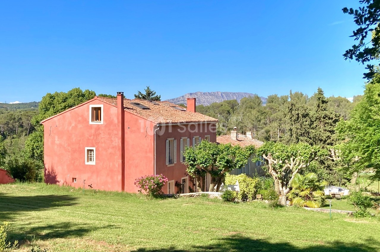 Maison provençale rouge entourée de verdure, avec un ciel bleu clair et des collines en arrière-plan.