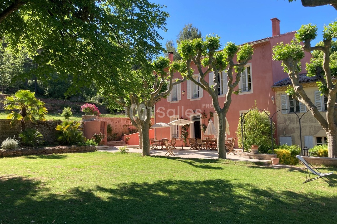 Façade d'une maison provençale rose entourée d'arbres, avec une terrasse aménagée et un jardin verdoyant.