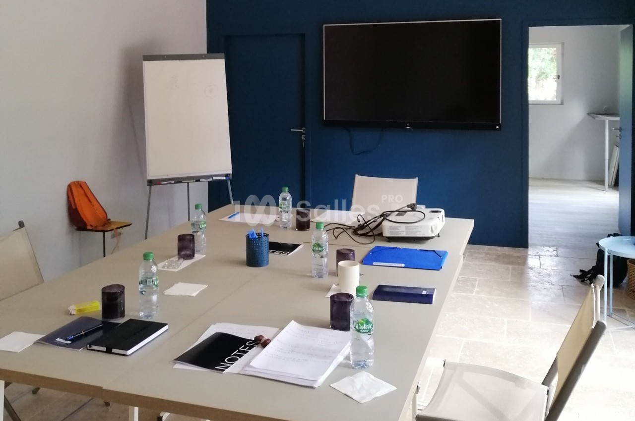 Salle de réunion avec table équipée de documents, bouteilles d'eau, un vidéoprojecteur et un écran mural.