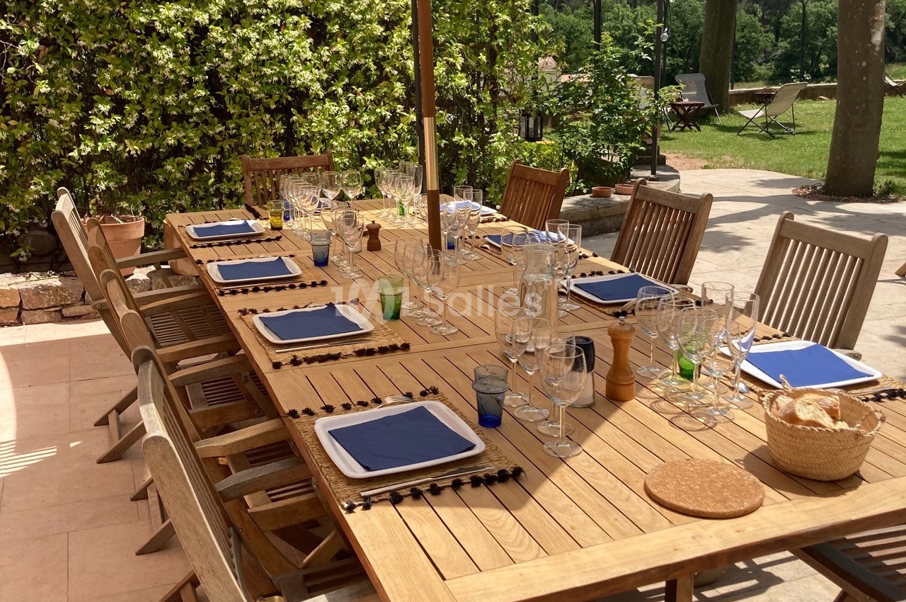Table en bois dressée pour un repas en extérieur, avec assiettes, verres et serviettes bleues, entourée de chaises.