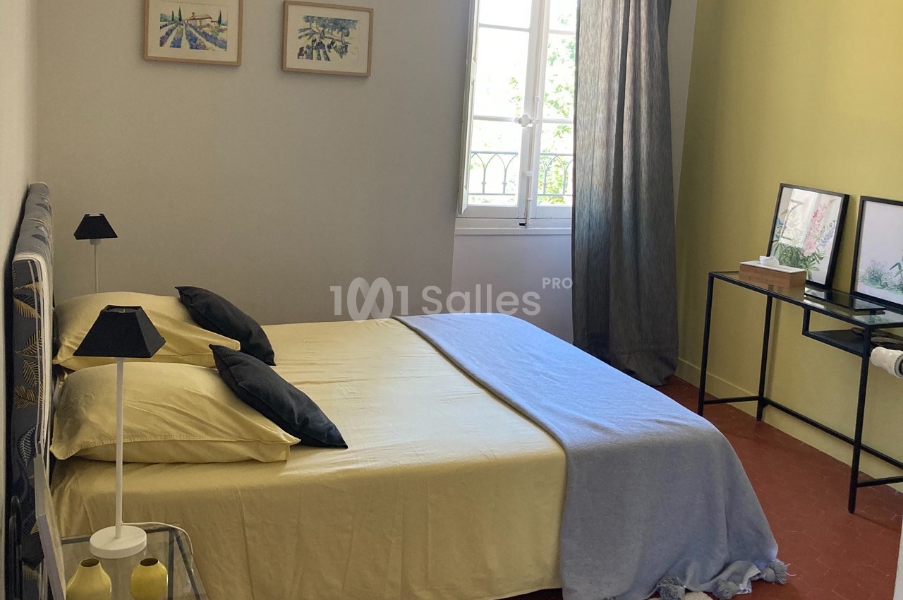 Chambre lumineuse avec lit double, draps jaunes, table de chevet, bureau et fenêtre donnant sur l'extérieur.