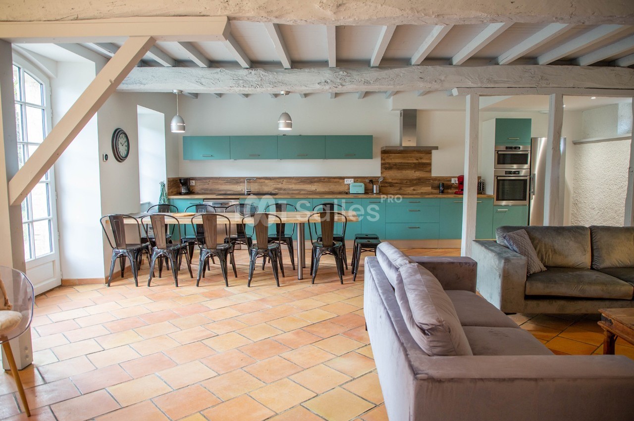 Cuisine ouverte avec meubles turquoise, grande table en bois entourée de chaises métalliques, et salon avec canapé gris.
