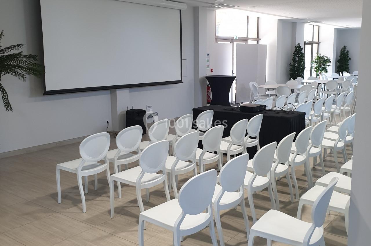 Salle de conférence lumineuse avec des rangées de chaises blanches alignées face à un écran de projection.