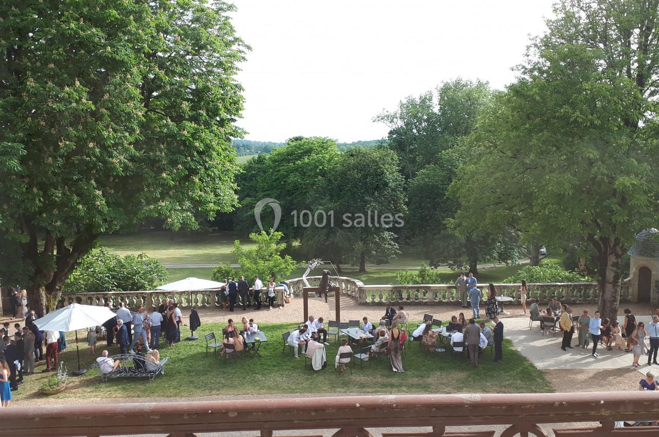 Groupe de personnes rassemblées dans un jardin arboré avec des tables, parasols et une vue sur un parc verdoyant.
