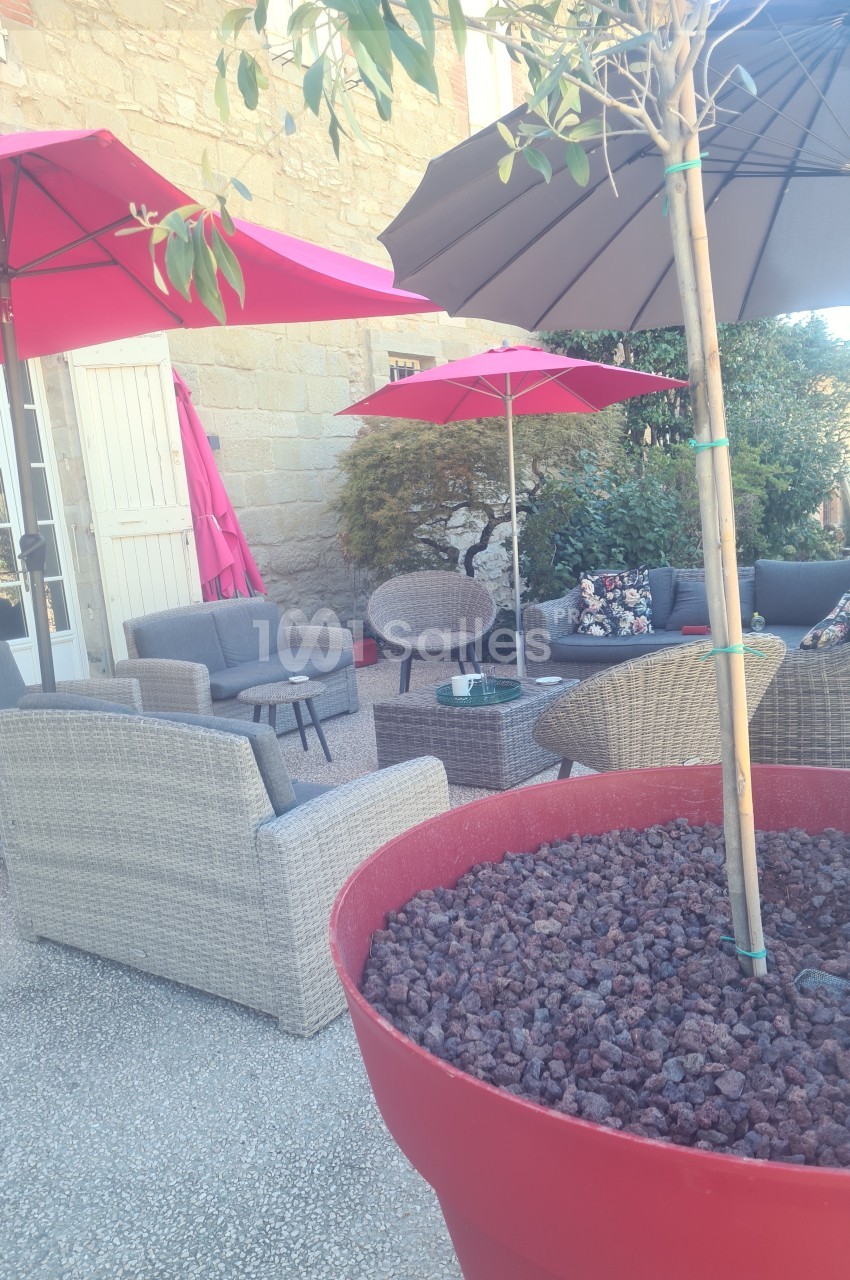 Salon de jardin avec fauteuils en rotin, parasols colorés et grande jardinière rouge sur une terrasse ensoleillée.