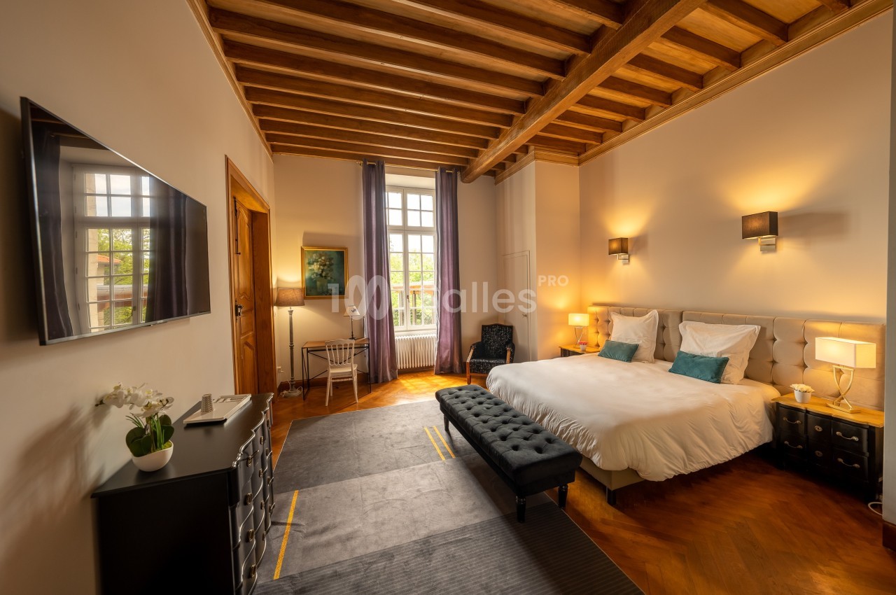 Chambre lumineuse avec lit double, poutres apparentes, bureau près de la fenêtre et décoration élégante.