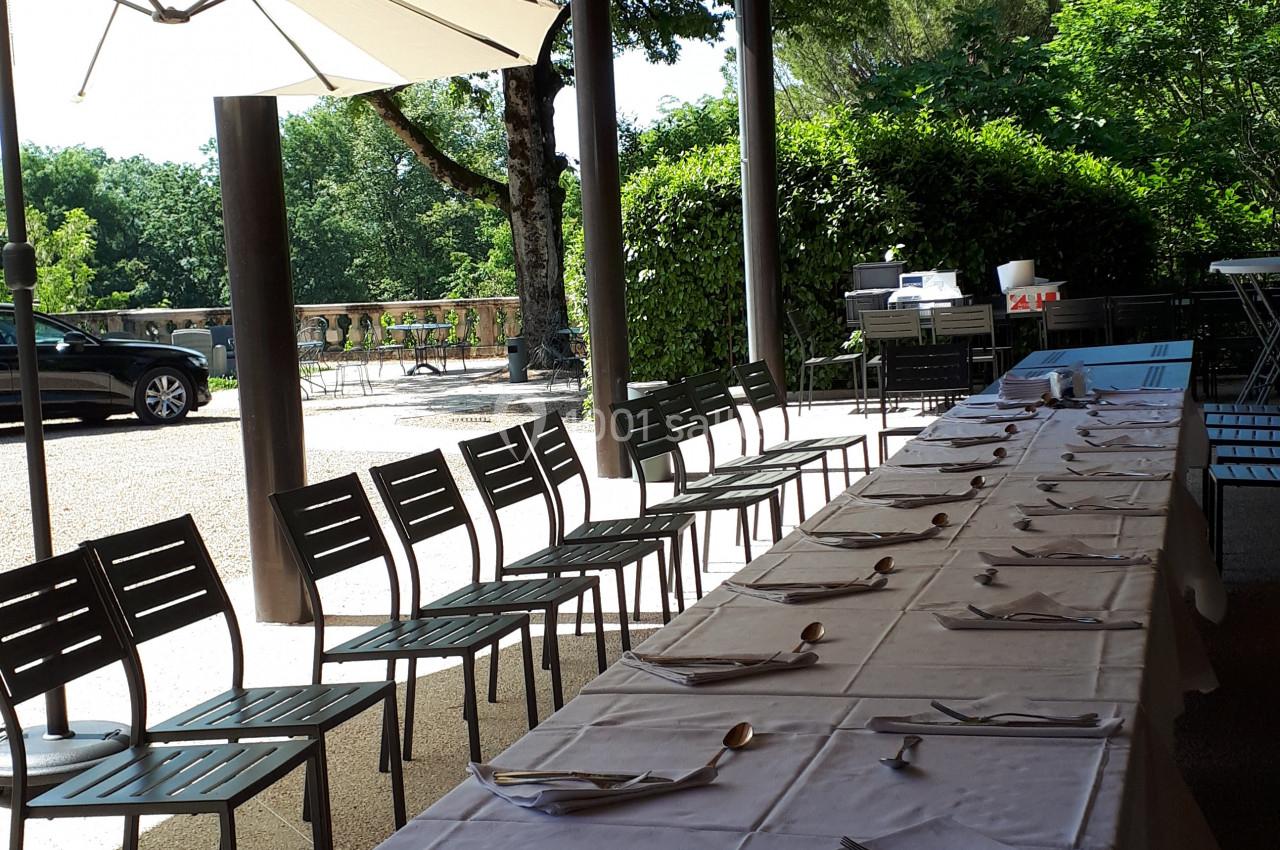 Tables alignées avec nappes blanches et couverts, disposées en extérieur sous une pergola en bois, entourées de chaises.
