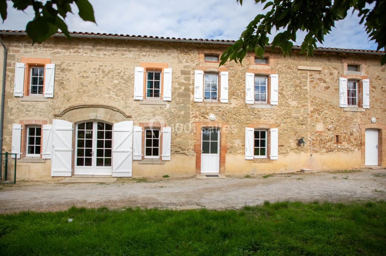 Façade d'une maison en pierre avec volets blancs, fenêtres à carreaux et une porte-fenêtre ouverte sur un jardin.