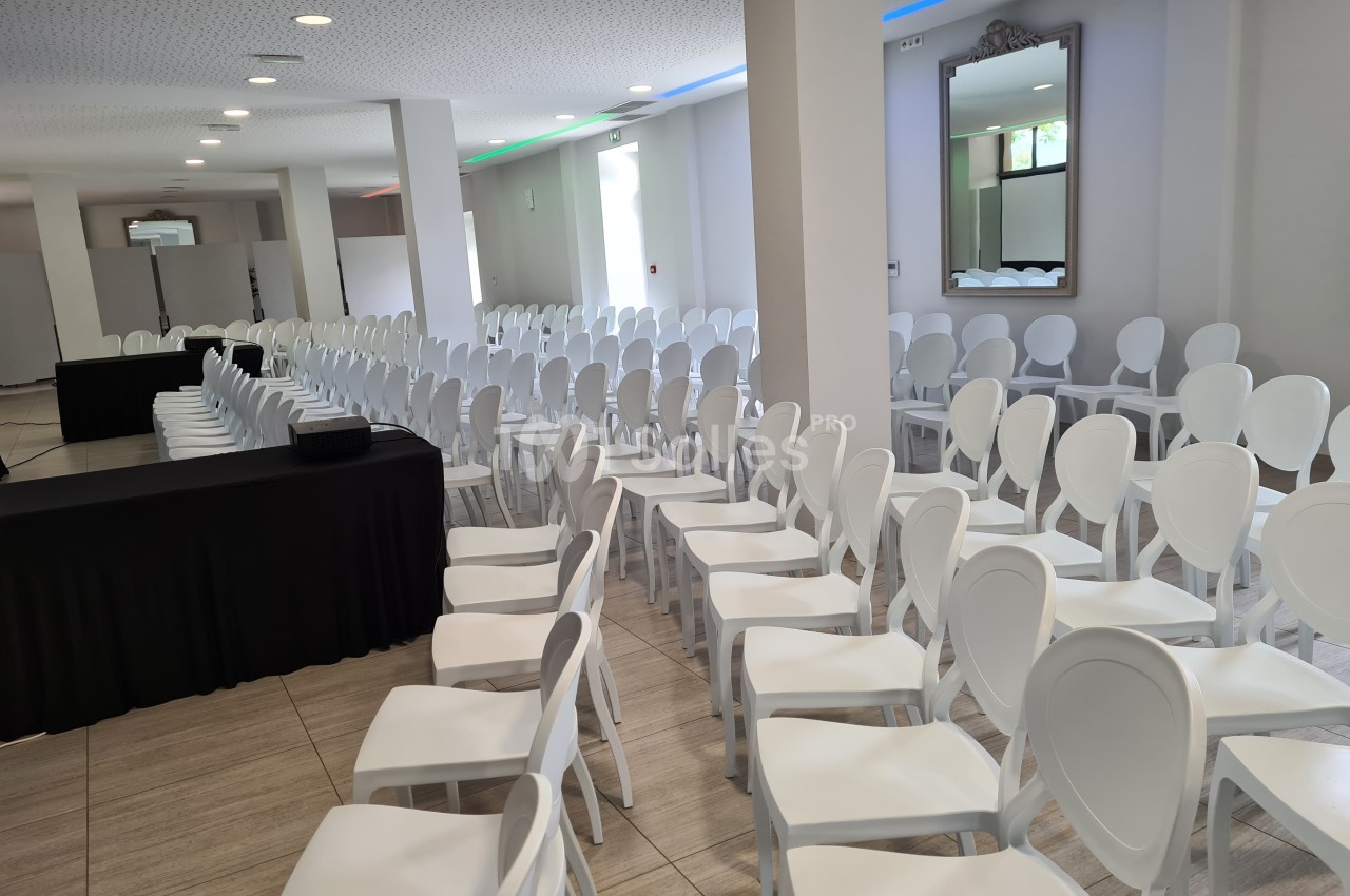Salle de conférence lumineuse avec des rangées de chaises blanches alignées face à une table noire.