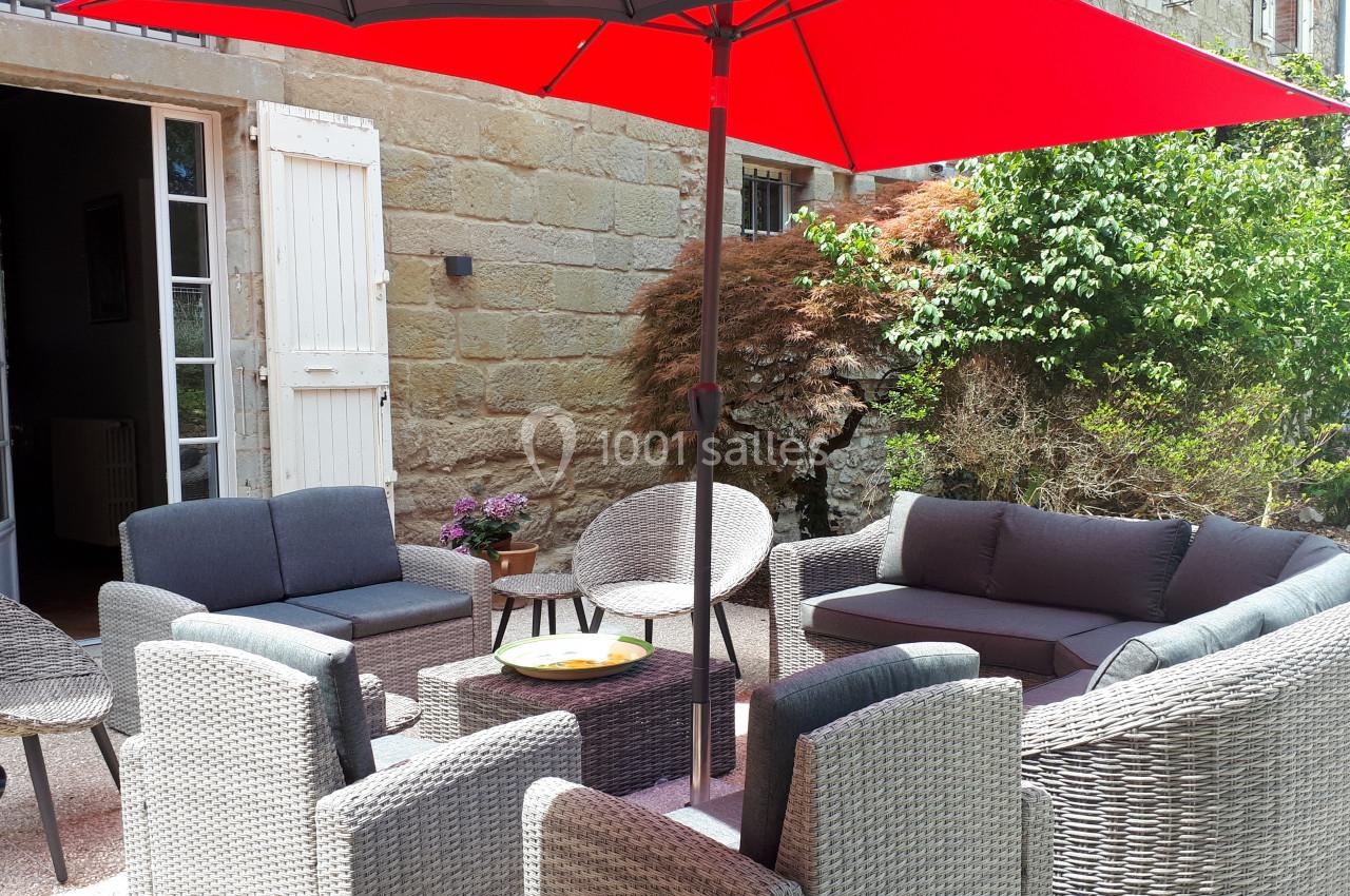 Salon de jardin en rotin avec coussins gris, disposé sous un parasol rouge, devant une maison en pierre.