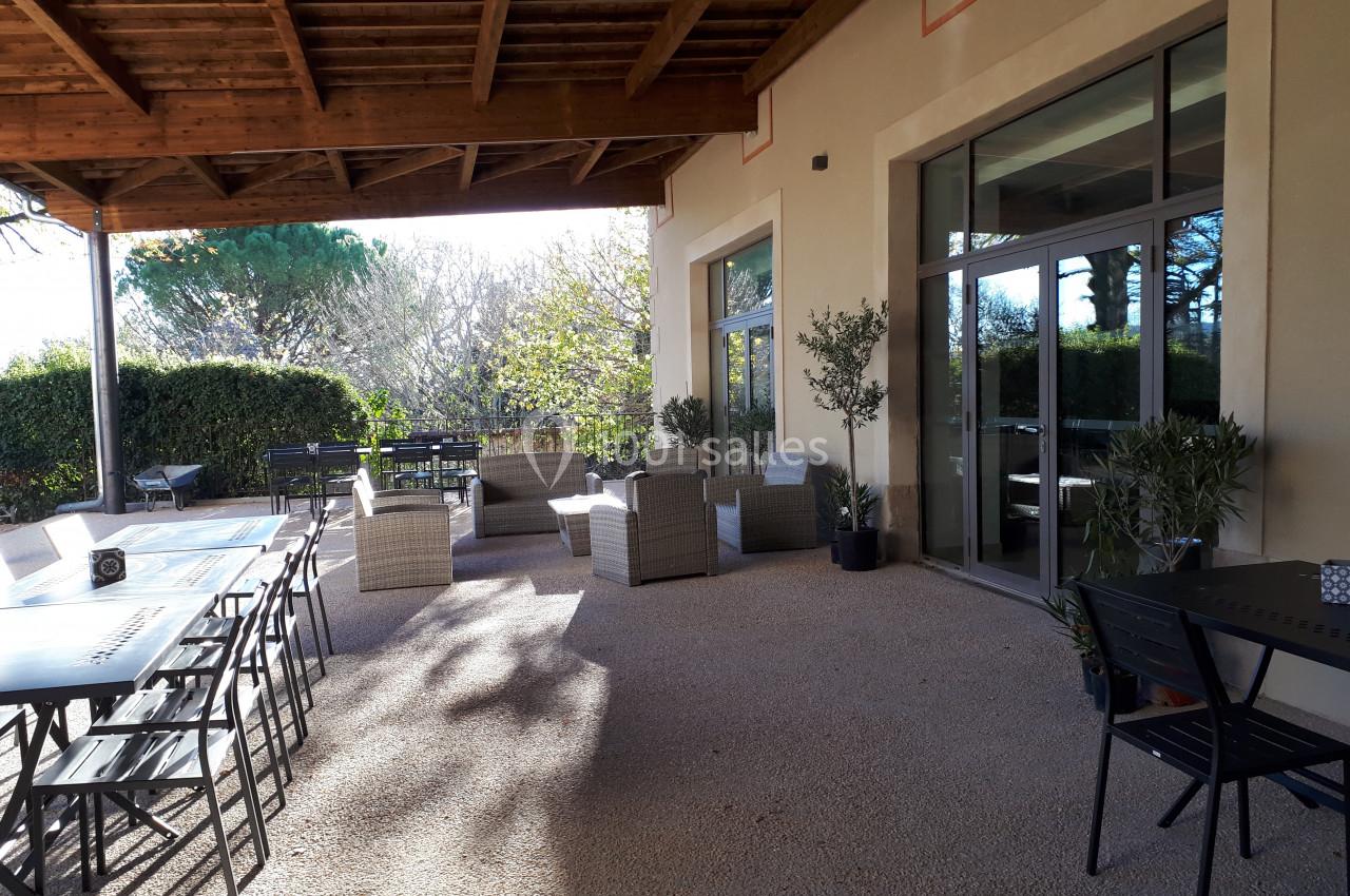 Terrasse couverte avec tables, chaises et fauteuils en rotin, entourée de verdure et baignée de lumière naturelle.