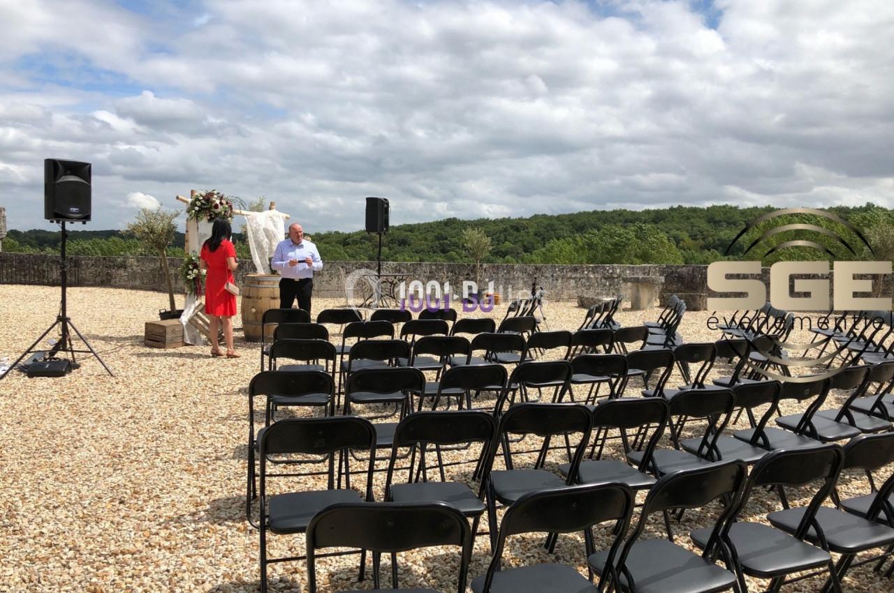 Chaises noires disposées en rangées sur un sol en gravier, avec une arche fleurie et des haut-parleurs en arrière-plan.